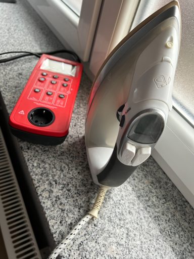 Gerätemessung nach DIN 0701-0702 (DGUV V3) Ein Bügeleisen und eine rote Steckdosenleiste auf einem Tisch neben einem Fenster.