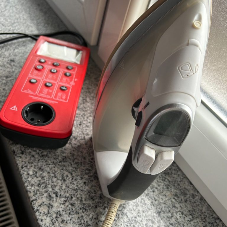 Geräteprüfung Ein Dampfbügeleisen neben einer roten Mehrfachsteckdose auf einer grauen Arbeitsplatte.