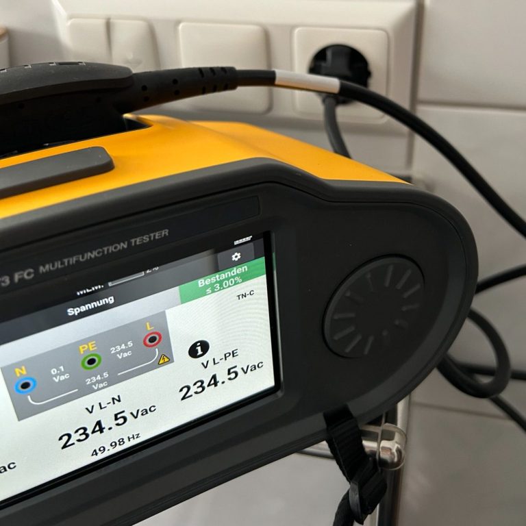 E-Check Ein gelbes Messgerät zeigt Spannung und Stromstärke an, angeschlossen an eine Steckdose.